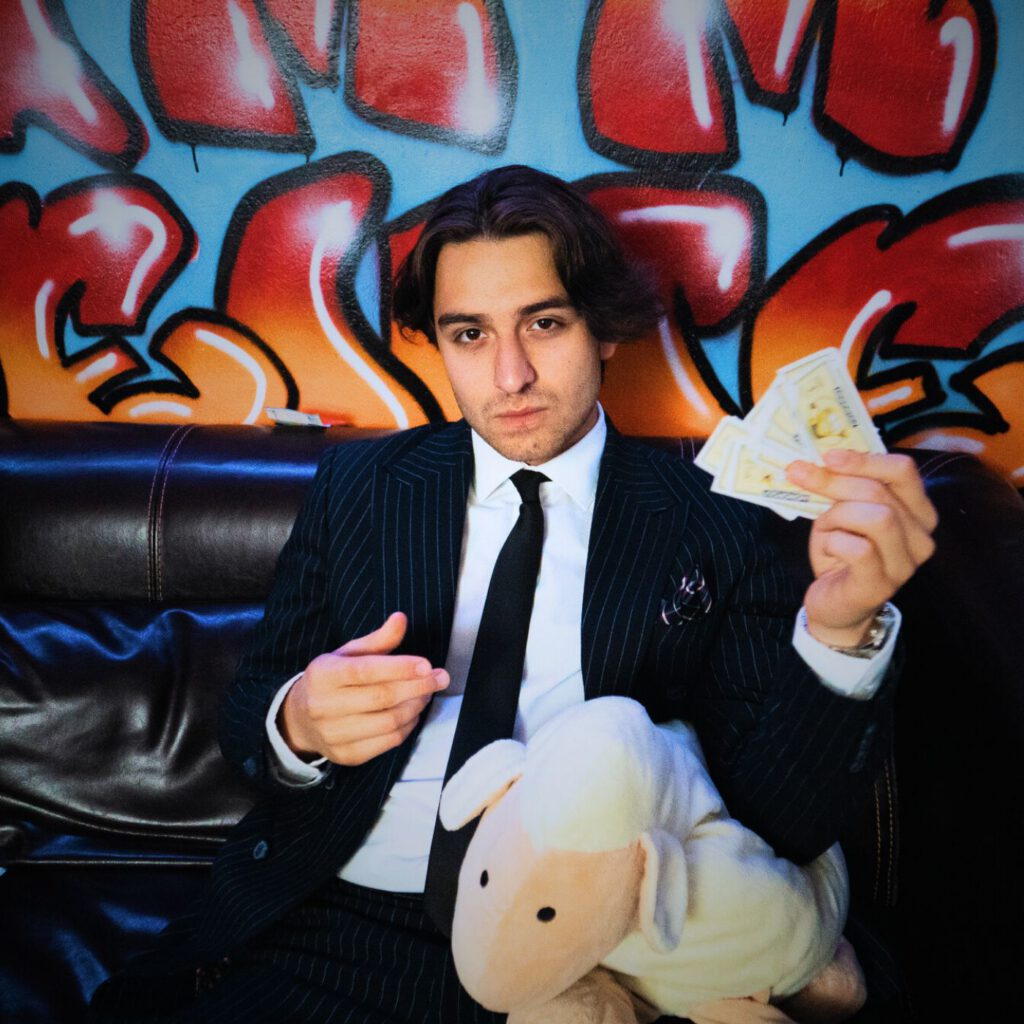 BaranBeatzz im Anzug mit Monopoly-Geld und Plüsch-Schaf vor der Graffiti-Wand im RNMS Studio, Cover zu „Besser als du“